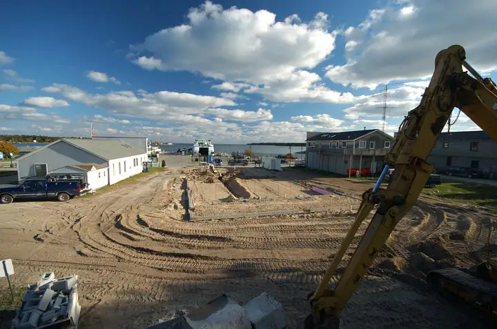 An elevated view shows a construction site with a rectangular foundation, excavator, buildings, and waterfront beyond.
