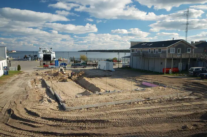 Construction site with a concrete foundation, heavy equipment, waterfront vessel, pier, and buildings.