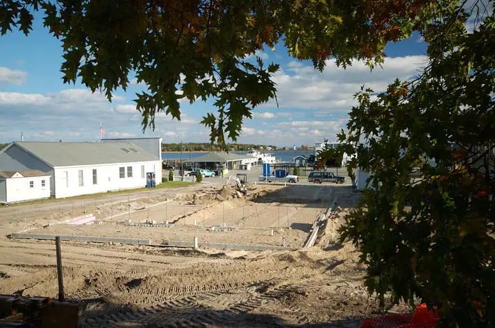A construction site with concrete foundation walls beside a white building and waterfront structures.