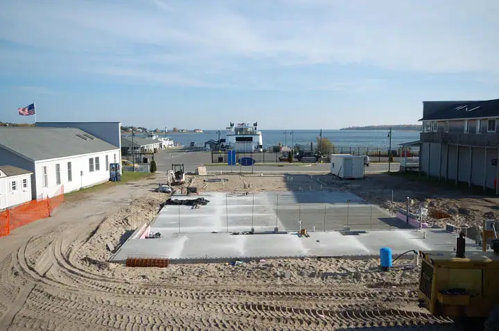 Concrete slab under construction beside a waterfront ferry terminal, with buildings, equipment, and tire tracks.