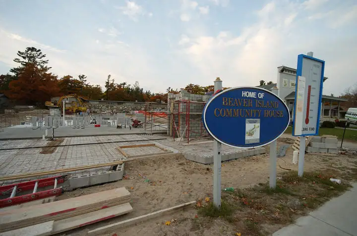 A blue community-house sign stands beside a concrete building under construction and a tall thermometer sign.