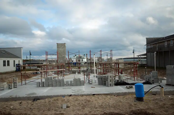 Construction site with concrete block walls, red scaffolding, a concrete slab, and a blue barrel beneath cloudy skies.
