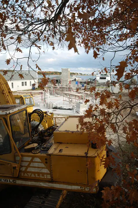 A yellow excavator overlooks a concrete-block construction site through autumn branches.