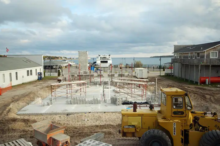 A concrete-block building foundation under construction beside a waterfront, with scaffolding and a yellow loader.