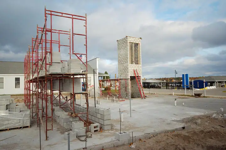 A concrete-block structure and red scaffolding stand on a broad construction site beneath a cloudy sky.