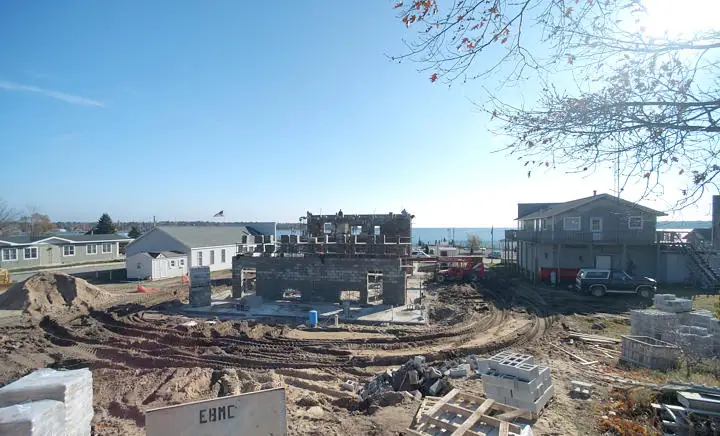 Construction site with exposed concrete-block walls, heavy equipment, nearby houses, and water visible beyond.