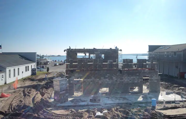 Concrete-block building under construction beside a waterfront, with scaffolding and nearby buildings.