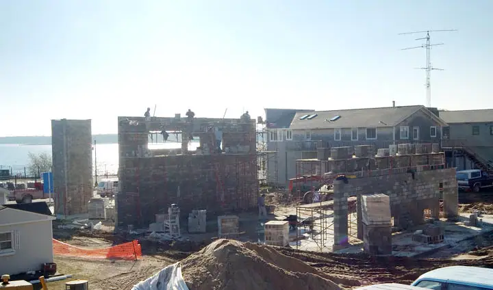 Concrete-block walls rise at a construction site beside existing buildings and a broad body of water.