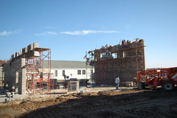 Concrete block walls rise around workers and scaffolding at a construction site beside an orange loader.