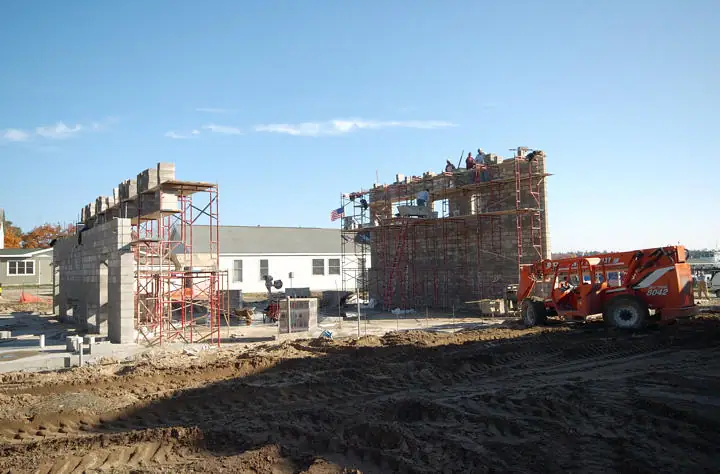 Concrete-block buildings rise behind red scaffolding at a muddy construction site, with an orange loader in the foreground.