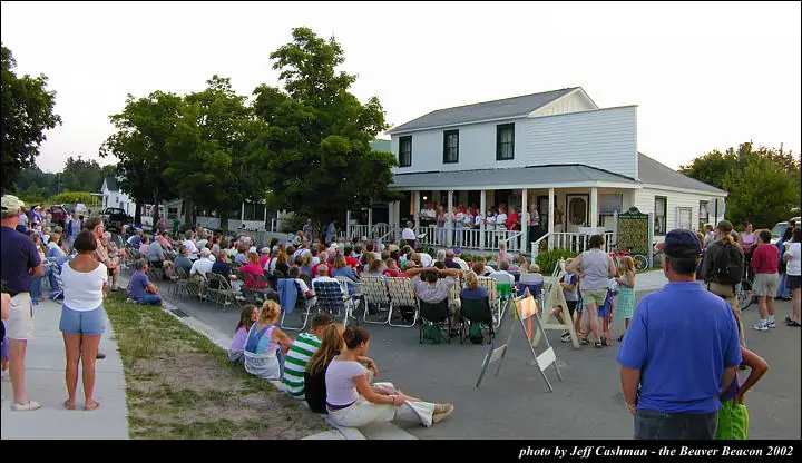 A large crowd sits in folding chairs and on the ground facing people gathered on a house porch.