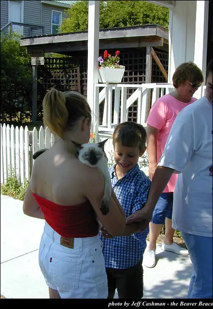 A woman holds a cat while a smiling boy reaches toward it beside a porch and picket fence.