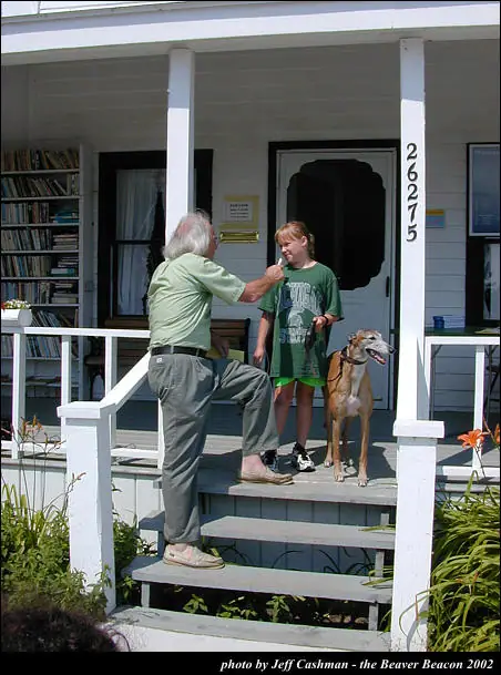 An older man, a young person, and a leashed dog stand on the steps of a house porch.