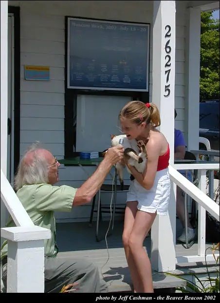 An older man uses electric clippers on a cat held by a young woman on a porch.