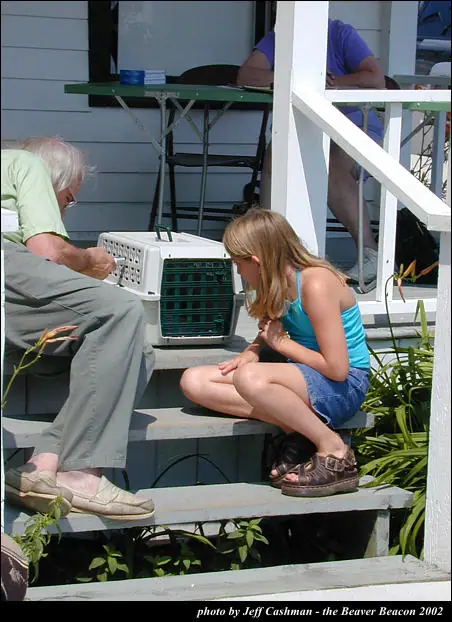 An older man works on a pet carrier while a girl crouches beside him on porch steps.