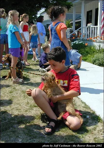 A boy sits on grass holding a small dog while children gather beside a porch and garden.
