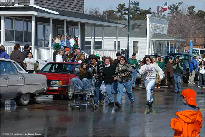 People in festive clothing run down a wet street as spectators gather outside storefronts.