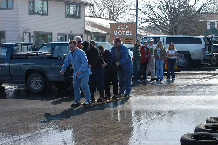 People balance together on a narrow wooden plank in a wet parking area while others watch.