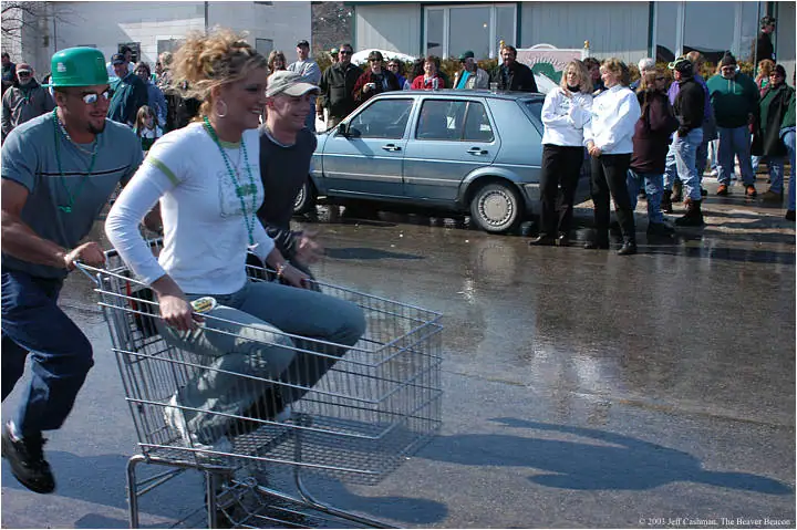 A woman rides in a shopping cart as a man pushes it past spectators gathered on a wet street.
