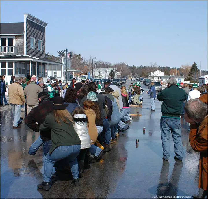 A crowd watches and takes part in a tug-of-war on a wet street lined with buildings and parked cars.