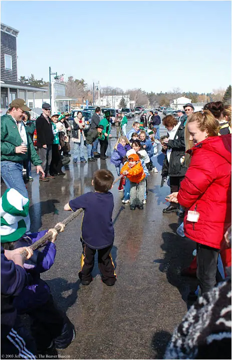 Children pull a rope in a tug-of-war while adults and spectators watch along a wet street.