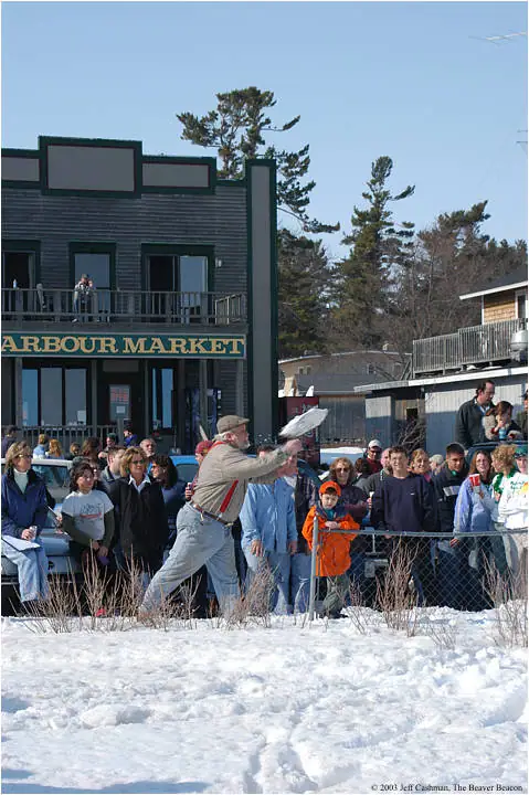 A man throws a white flying disc before a crowd beside a storefront in the snow.