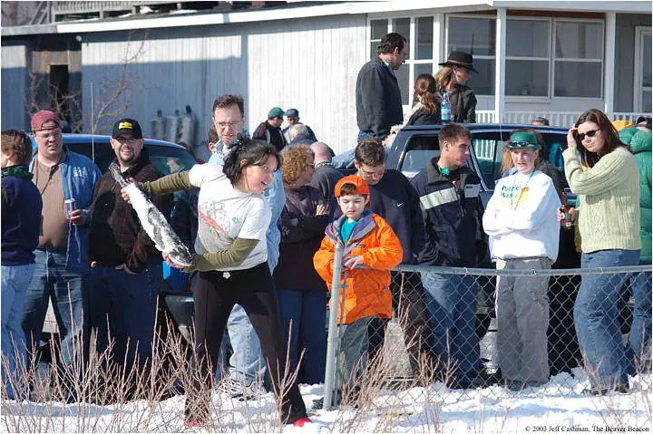 Adults and children gather in snow behind a chain-link fence as a woman holds a long silvery object.