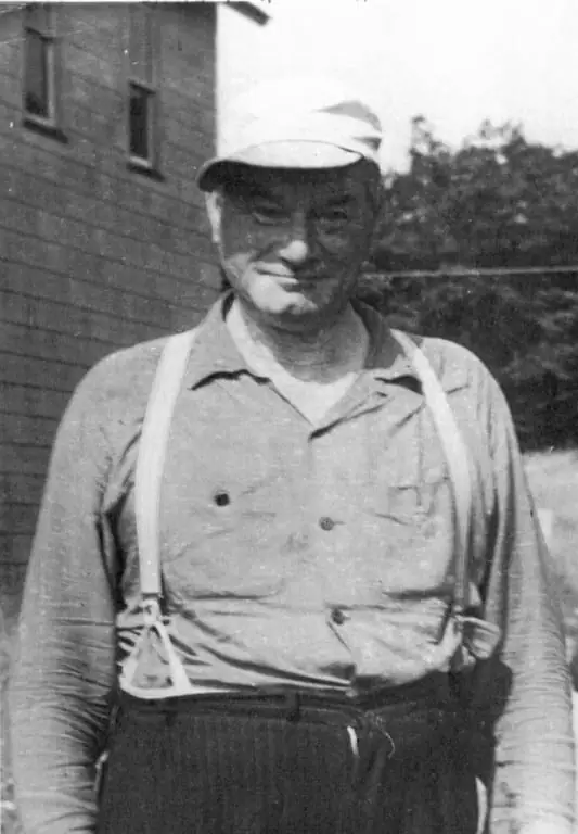Black-and-white portrait of a man in a cap and suspenders standing beside a wood-sided building.