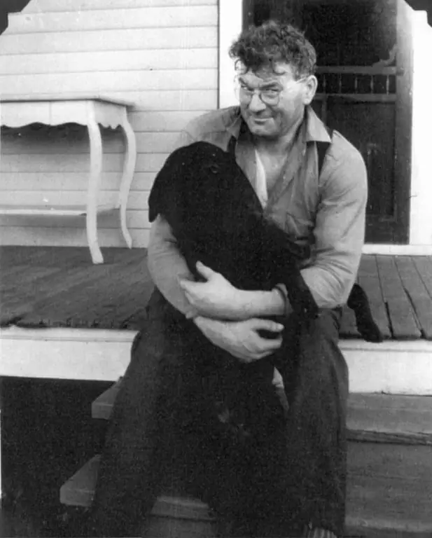 A man in glasses sits on a wooden porch while holding a large black dog.