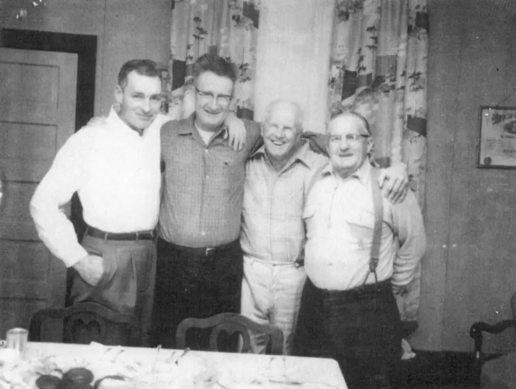 Four smiling men stand arm in arm indoors behind a table with chairs in the foreground.