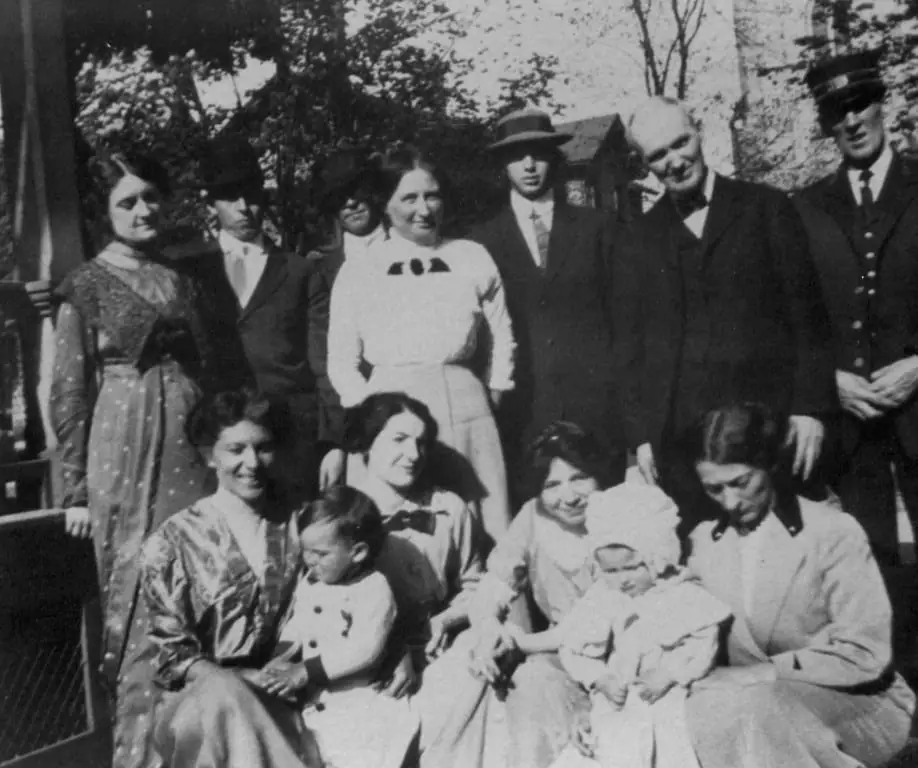 A large group of adults and two young children posed outdoors in front of trees and a building.