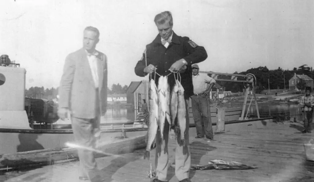 A man displays four fish on a dock while other people stand nearby and boats and waterfront buildings appear behind him.