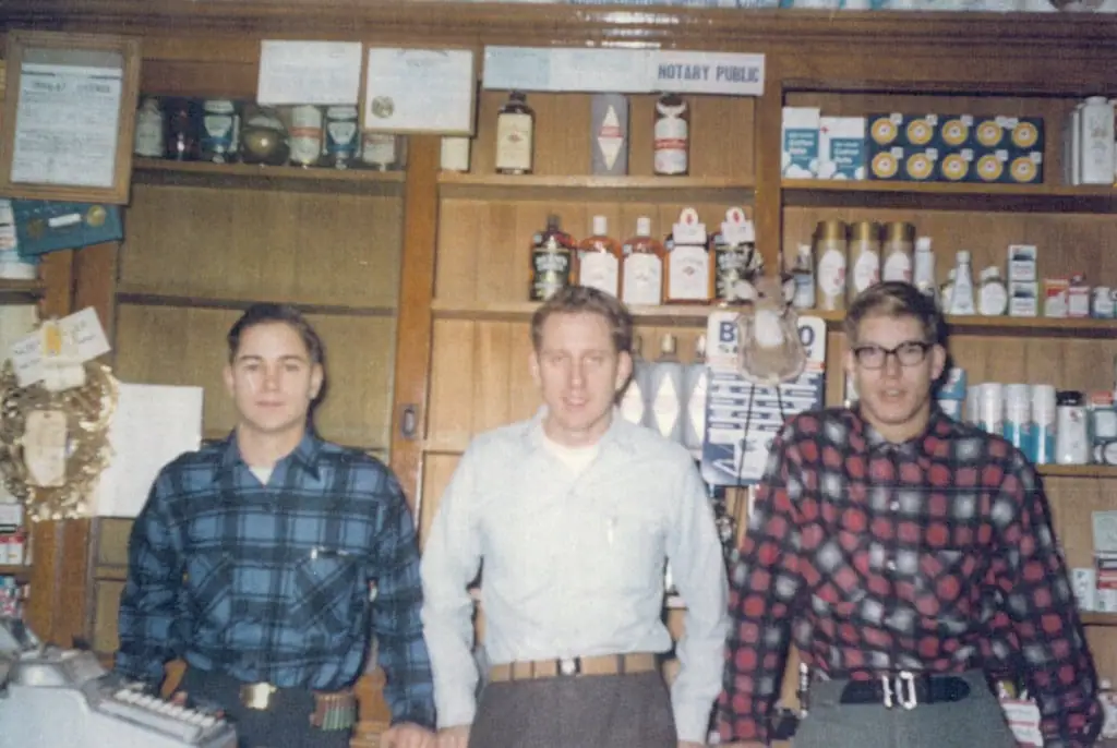 Three men stand in front of shelves stocked with bottles, boxes, and other merchandise inside a shop.