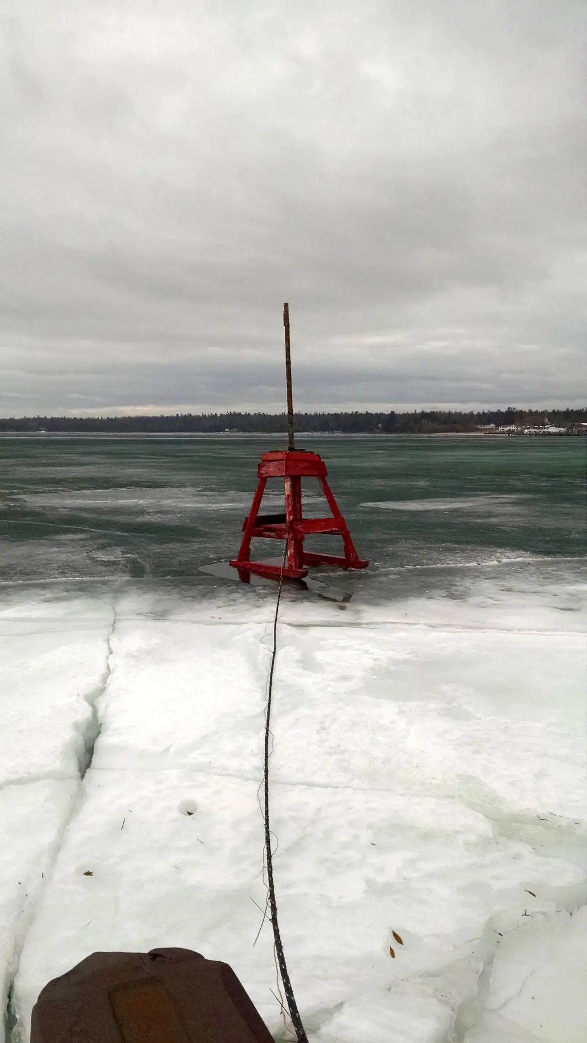 A red wooden buoy stands amid frozen water, with a rope extending across the ice.