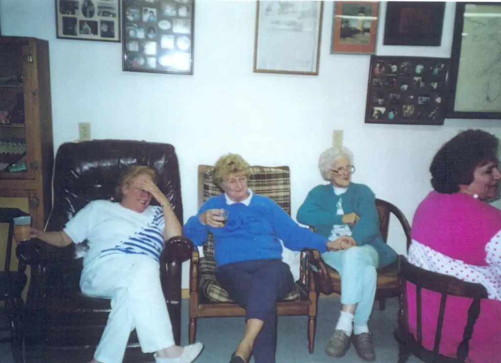 Four women sit in armchairs indoors, smiling and holding drinks.