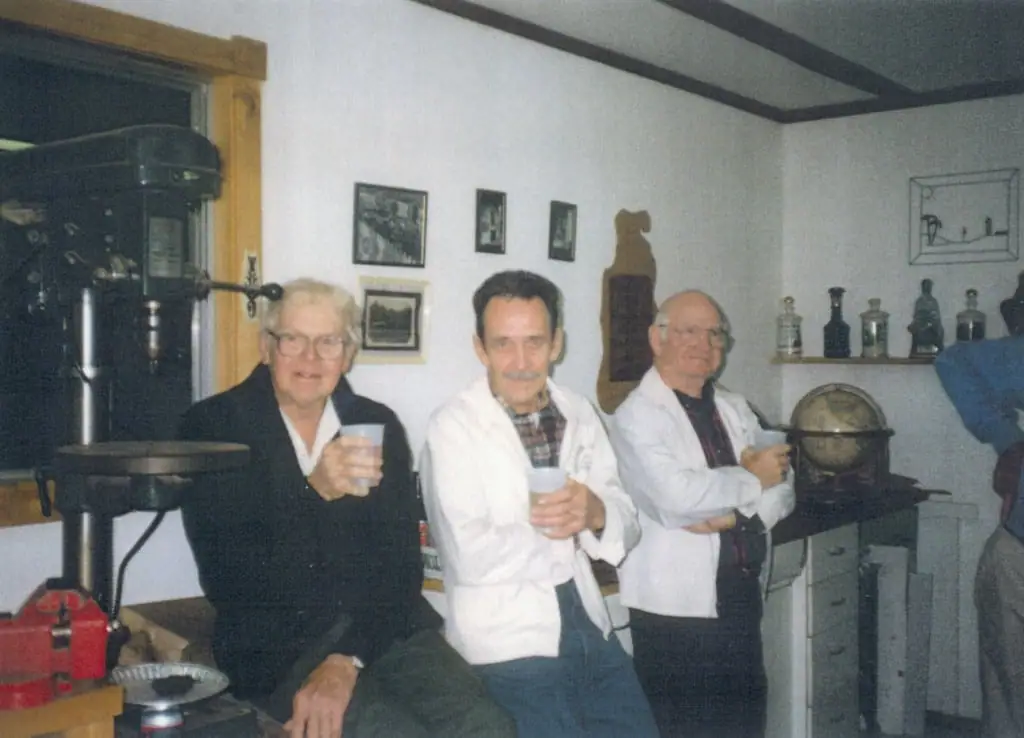 Three men in white coats and a dark jacket pose with cups inside a room containing equipment, bottles, and a globe.