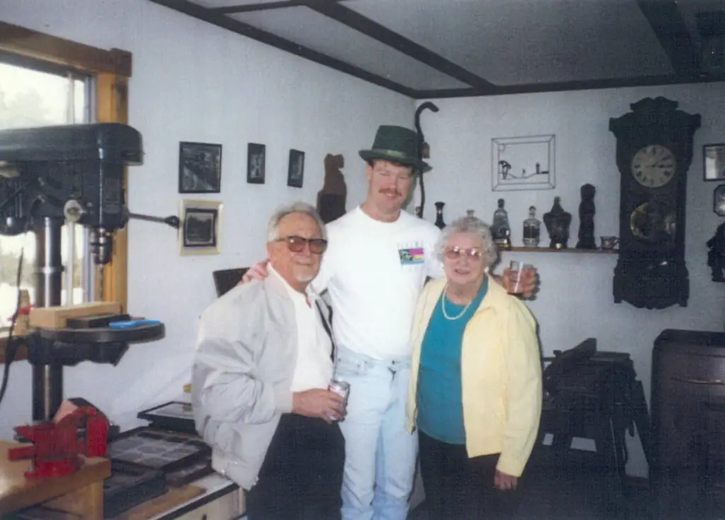 Three people pose together in a room containing a drill press, framed pictures, bottles, and a grandfather clock.