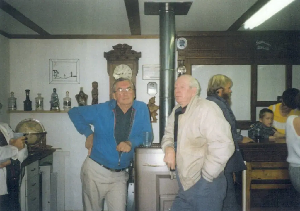 People gather indoors around a large stove, with a clock, shelves of objects, and a child visible in the room.