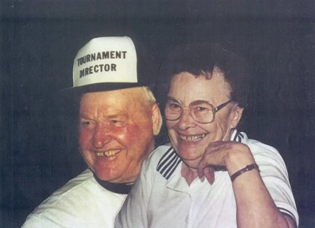 Two smiling adults pose together; the man wears a cap labeled “TOURNAMENT DIRECTOR.”