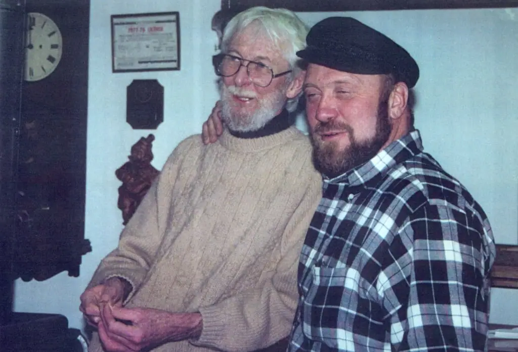 Two bearded men pose together indoors, one wearing glasses and a sweater and the other a cap and plaid shirt.