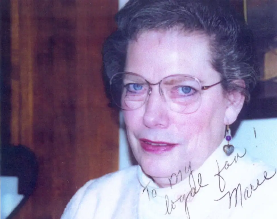 Close-up portrait of a smiling woman wearing glasses and a heart-shaped earring, with a handwritten dedication across the lower image.