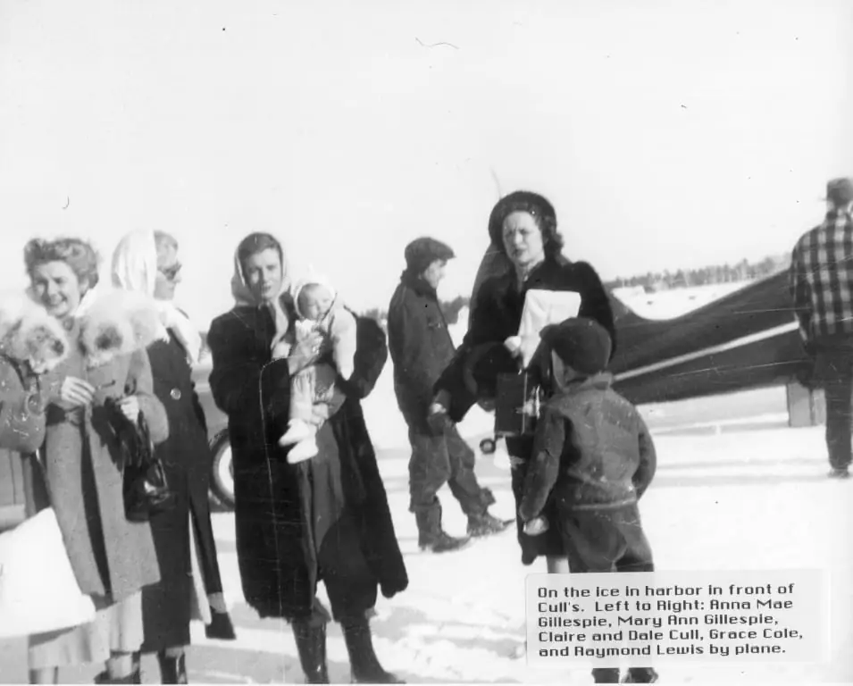 People stand on a snow-covered frozen surface beside a small airplane, with two adults holding babies.