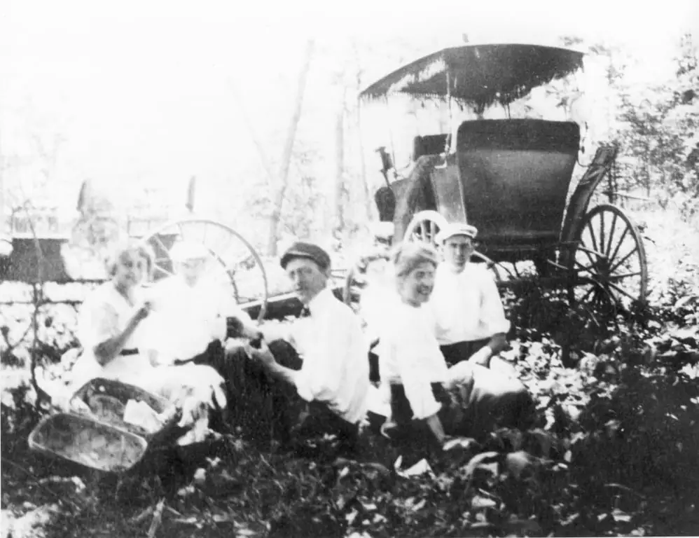 Five people pose outdoors beside a large covered horse-drawn carriage in a black-and-white photograph.