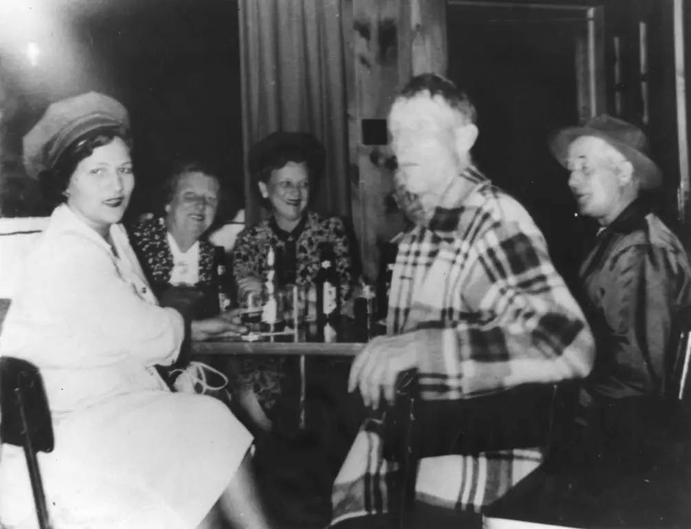 Five adults sit around a table in a wood-paneled room, with bottles and glasses on the table.