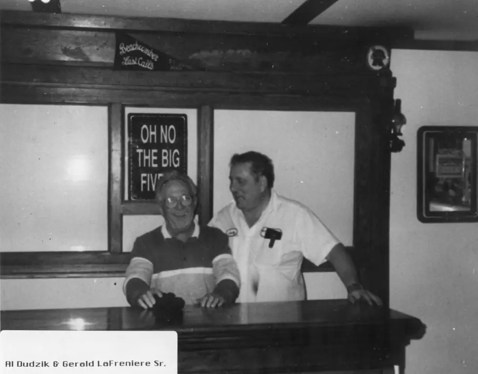 Two men stand behind a wooden bar beneath signs and a framed photograph caption.