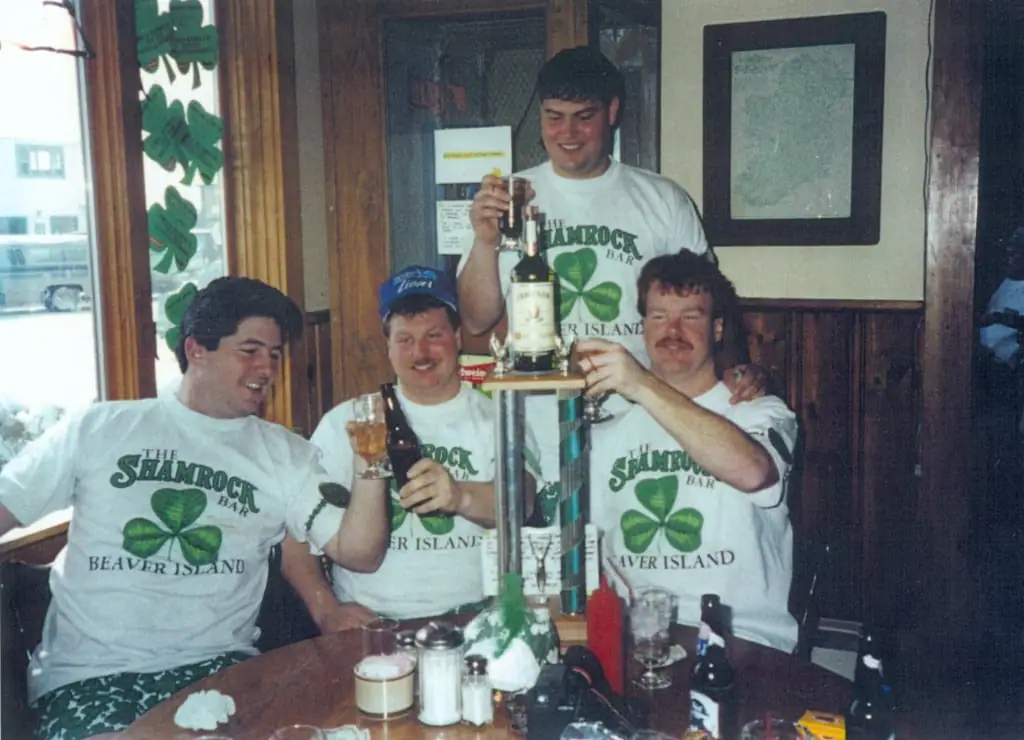 Four men in matching shamrock shirts gather around a table with bottles and glasses inside a wood-paneled bar.