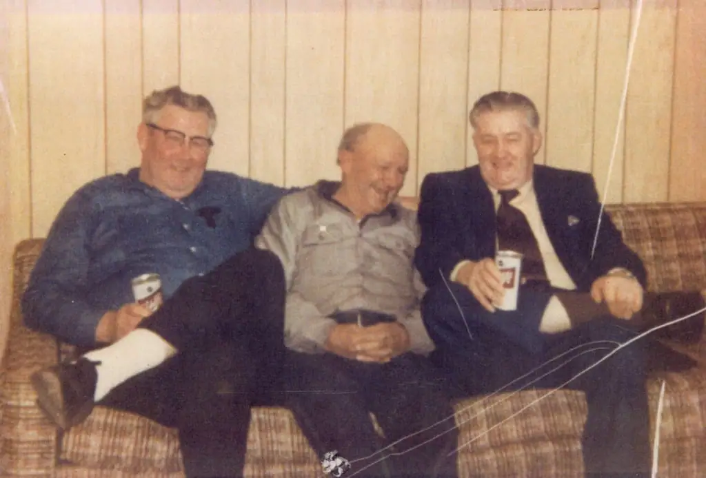 Three men sit together on a patterned couch, smiling and holding paper cups.