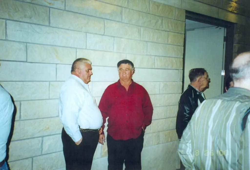 Adults stand beside a light-colored stone wall and doorway, with other people partly visible at the edges.
