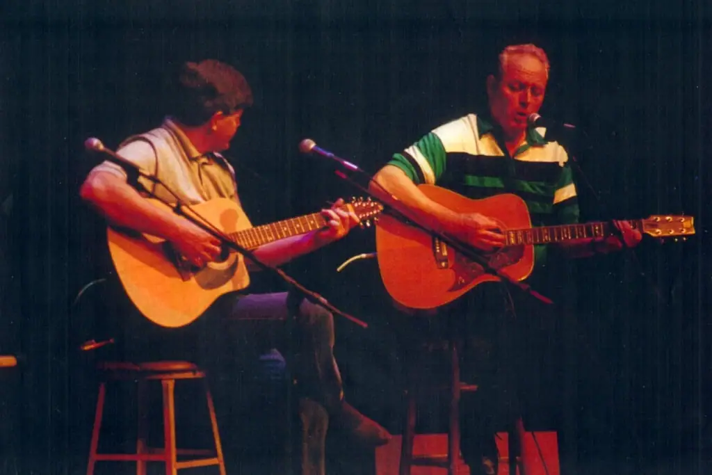 Two seated musicians play acoustic guitars on a dimly lit stage.