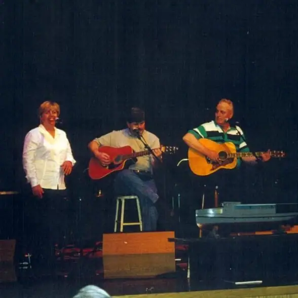 Three musicians perform on a dimly lit stage, with two acoustic guitars and a woman standing beside them.